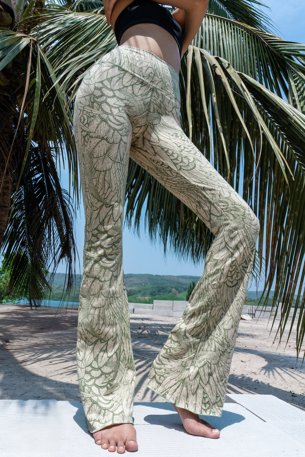 Model wearing Funky Simplicity Flares in Cream Olive Feather print made from cotton lycra - with green palm trees and a river on the background - shown from the front