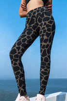 Woman wearing Funky Simplicity Giraffe Leggings in the colour Olive Black shown from the front with an ocean and blue sky on the background
