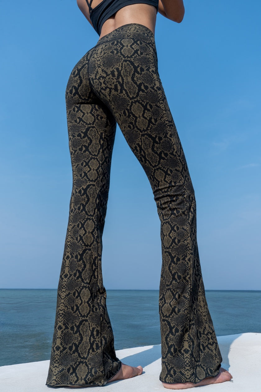 Woman wearing olive black Snake patterned flared leggings shown from the back, from Funky Simplicity - standing on a white surface with a blue sky and ocean background