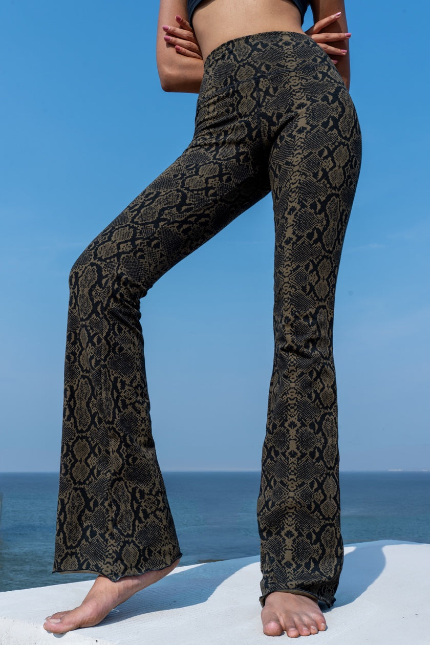 Woman wearing olive black Snake patterned flared leggings from Funky Simplicity - standing on a white surface with a blue sky and ocean background