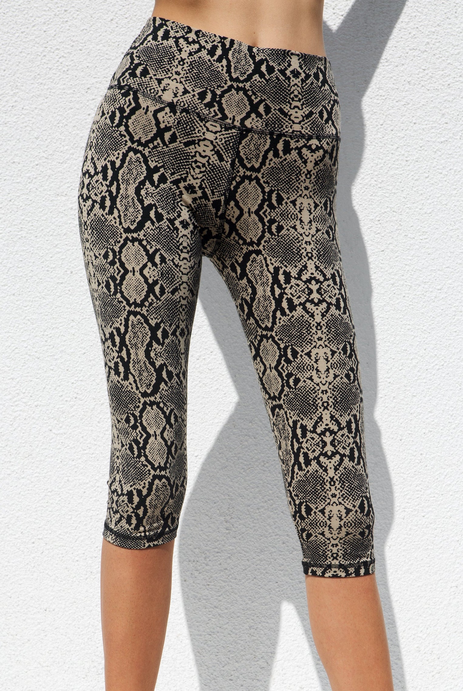 A model wearing high-waisted cream-colored capri leggings with a black snake print, standing against a white background.