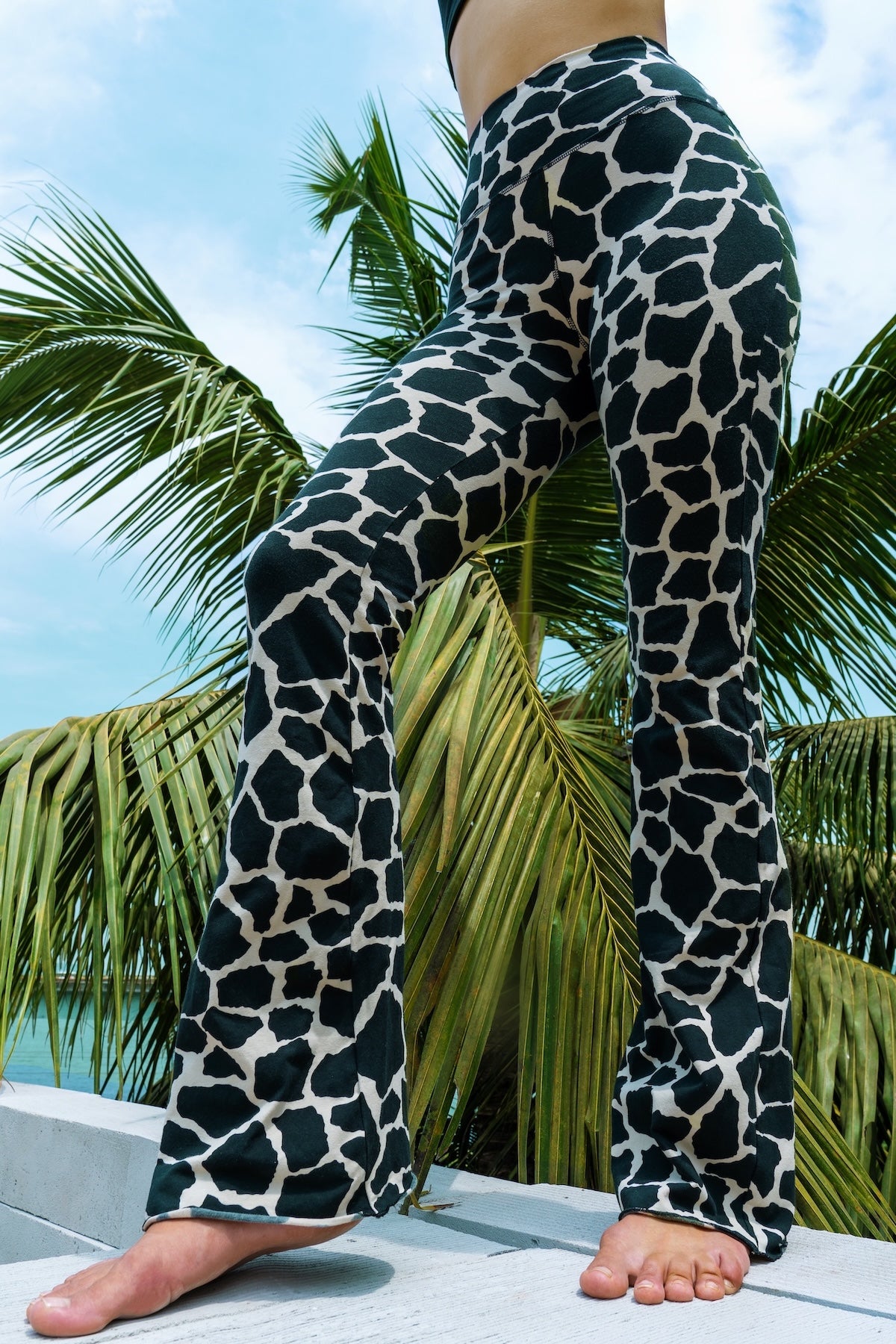 Woman wearing cotton lycra Flares in Cream Black Dutch Cow print from Funky Simplicity shown from the front with  leaves of a palmtree in the background with a cloudy blue sky